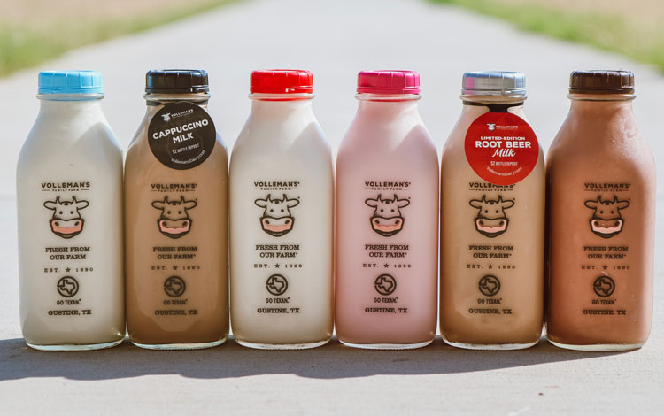 Seis botellas de vidrio llenas de varios sabores de leche se alinean en una acera que exhibe el producto lácteo de Volleman's Dairy.